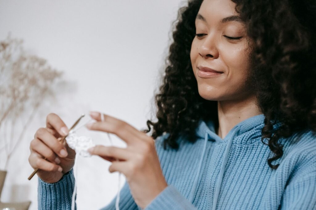 A smiling woman in a blue hoodie crocheting with a hook, indoors.
